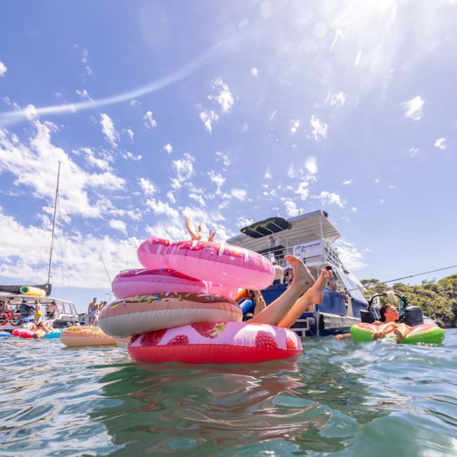Individuals on floats and inflatable rings enjoy a sunny day on the water near boats, under a clear blue sky, with the vibrant tunes of a DJ boat hire Sydney creating the perfect backdrop.