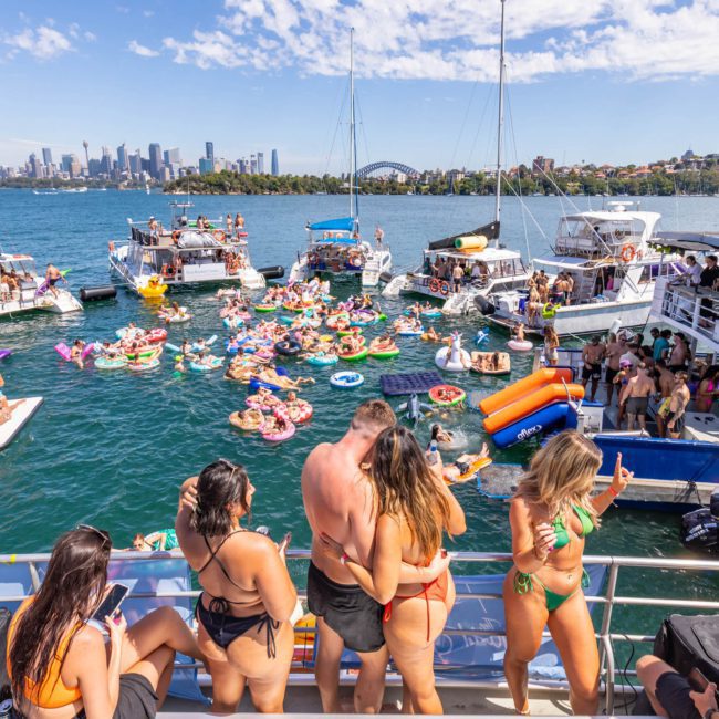 People on boats and in the water enjoying a sunny day, with many on inflatable floats. The background features a city skyline under a clear sky, perfect for a Sydney boat party hire.