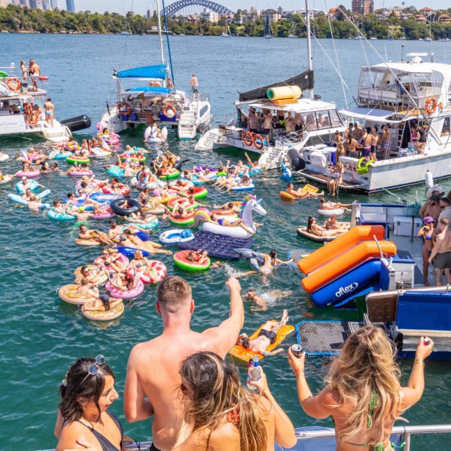 A group of people on boats and inflatable floats gather for a Sydney boat party hire with a cityscape and bridge in the background.