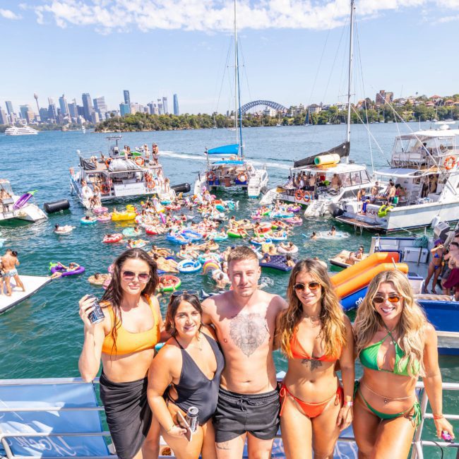 A group of five people in swimwear stands on a boat in front of a crowded marina with numerous boats and inflatables in the water. The city skyline and a bridge are visible in the background, creating the perfect setting for a Sydney boat party hire.