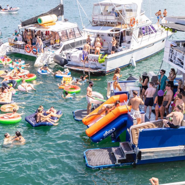 A crowded scene of people on boats and inflatables enjoying a sunny day on the water, with some individuals using water slides from the boats. Buildings are visible in the background, highlighting what a Catamaran party Sydney experience looks like.