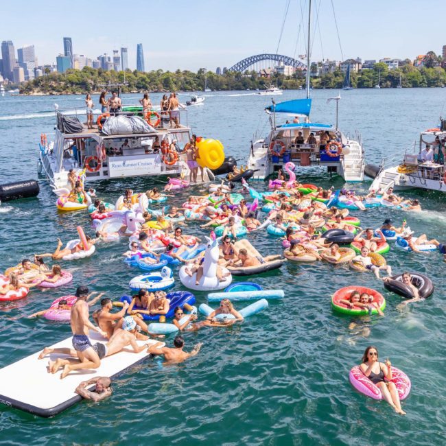 A large group of people on colorful inflatables gather between boats on a sunny day in a harbor, with a cityscape and a prominent bridge visible in the background, enjoying the vibrant atmosphere of a catamaran party Sydney.