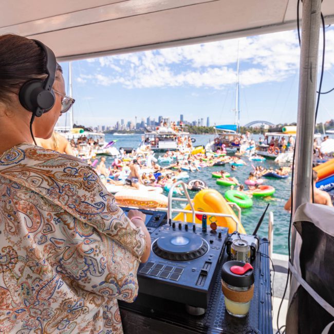 A DJ in patterned clothing is performing on a private yacht charter in Sydney Harbour while a large crowd on inflatable floats and boats enjoys a sunny day on the water.