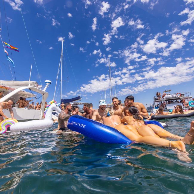 A group of people having fun in the water by several boats, with some on inflatable floats under a sunny sky with scattered clouds. Perfect for a Sydney boat party hire or private yacht charter on Sydney Harbour.