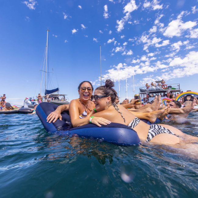 People relaxing on inflatables in the water near anchored boats on a sunny day, enjoying the vibe of a DJ boat hire Sydney.