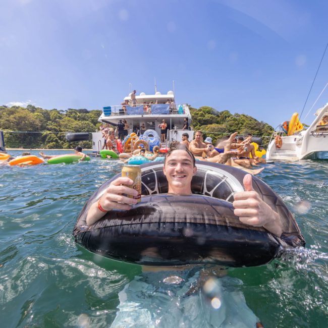 Person floating in the water on an inner tube, holding a drink and giving a thumbs-up, with boats and several people in the background enjoying a sunny day during a vibrant catamaran party in Sydney.