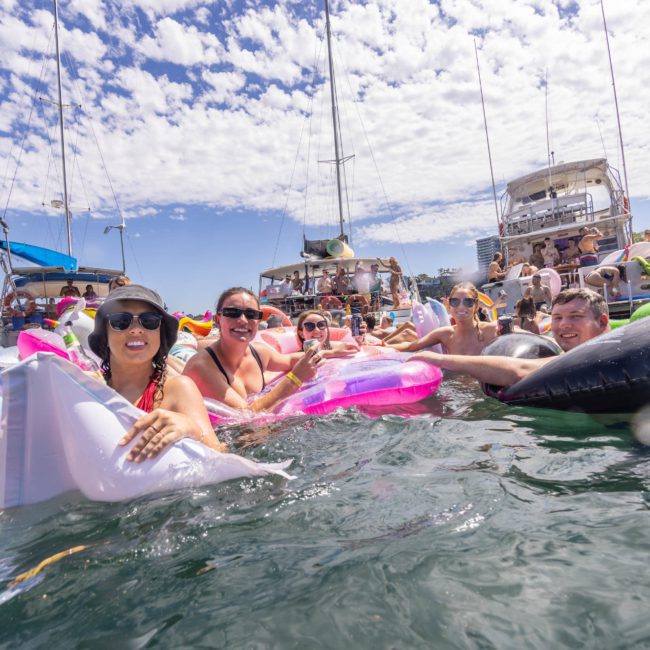 A group of people in swimsuits is floating on various inflatables in the water near some boats. The sky is partly cloudy, and a tree is visible in the background, creating a perfect setting for enjoying a catamaran party in Sydney.