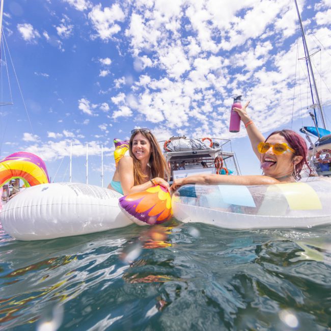 Two people are floating on an inflatable raft in a body of water, surrounded by boats. One person holds a drink and both appear to be enjoying the sunny day, perhaps after a fun DJ boat hire Sydney experience.