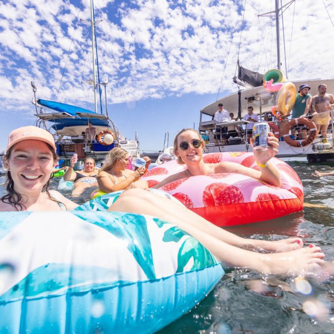 People enjoying a sunny day on inflatable floats in the water near anchored boats, with others socializing on board, epitomizing a perfect Catamaran party in Sydney.