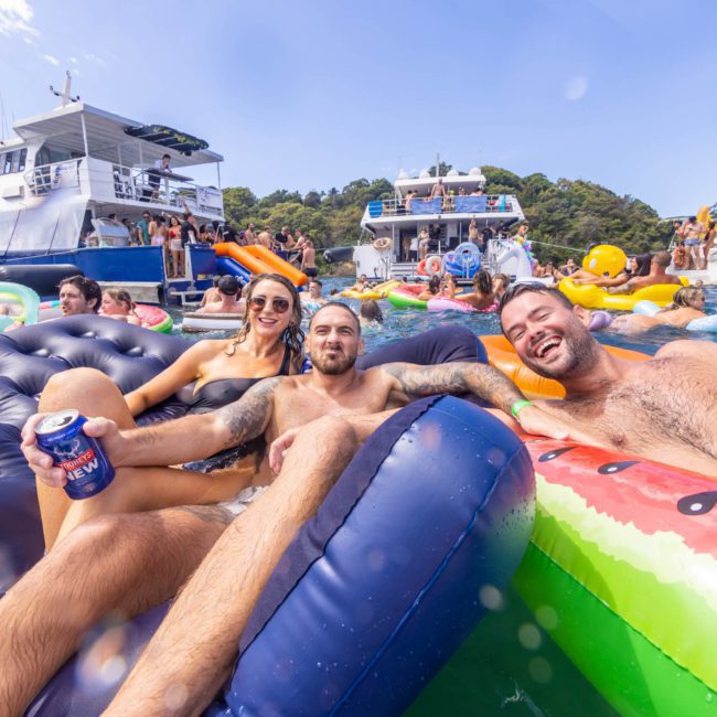 Three people relax on inflatable loungers in the water, holding drinks. In the background, boats with other revelers create a lively atmosphere perfect for a Sydney boat party hire.