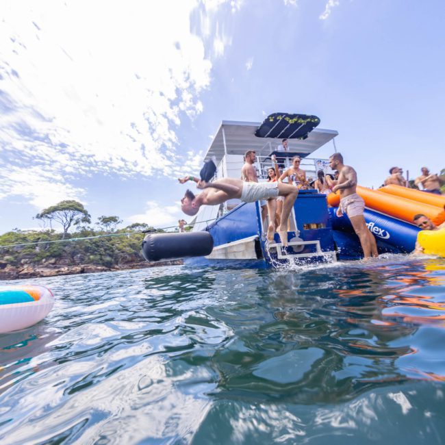 People on a catamaran party in Sydney jump into the water, while others enjoy slides and inflatables, surrounded by lush greenery and blue skies.