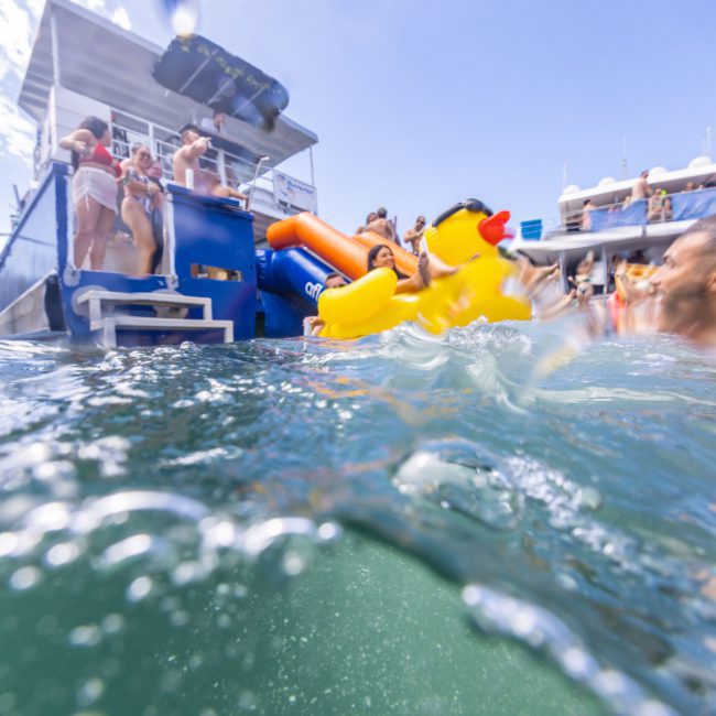 People in swimsuits enjoying a sunny day on a luxury yacht hire Sydney, with some in the water holding a large inflatable yellow duck.