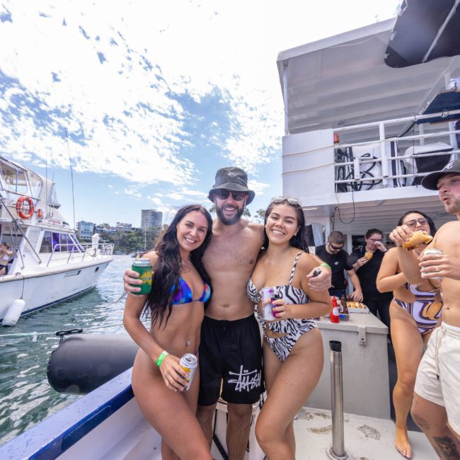 A group of smiling people in swimwear stand on a boat under a partly cloudy sky. Two people hold drinks while another person is eating. Other boats and buildings are in the background, setting the perfect scene for a memorable Sydney boat party hire.