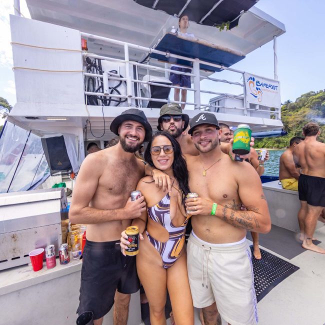 A group of young people in swimwear enjoys a sunny day on a catamaran party in Sydney with drinks in hand. The boat is at a scenic location with trees and water visible.
