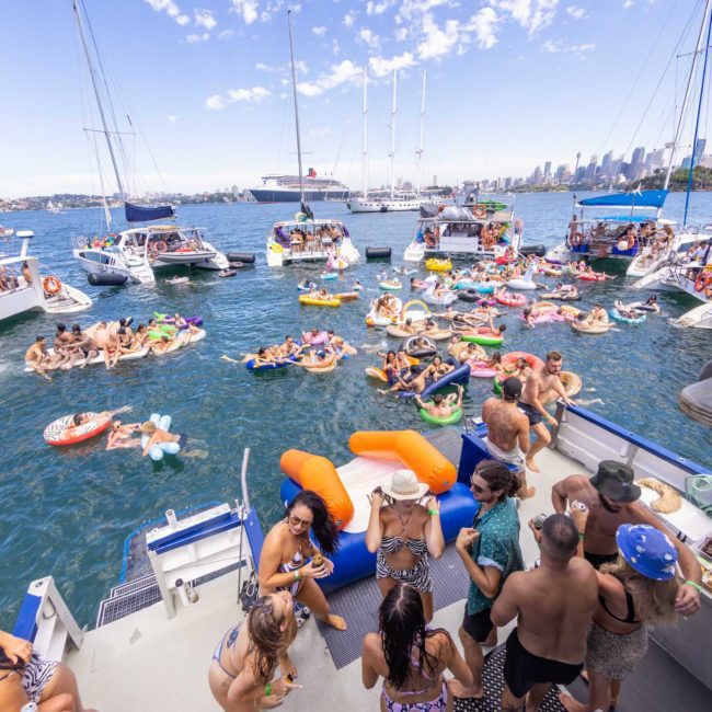 Boats are anchored close together in a bay with many people enjoying the water. Some are on inflatable floats and others are socializing on the boats. The city skyline is visible in the background, setting a perfect scene for a catamaran party Sydney or private yacht charter Sydney Harbour.