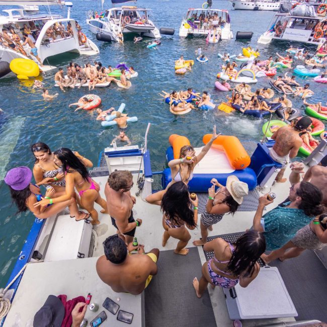 A large group of people are enjoying a sunny day on boats and inflatables in a crowded body of water, engaging in various activities like socializing and swimming. For an unforgettable experience, consider Sydney boat party hire or even luxury yacht hire Sydney for your next adventure.