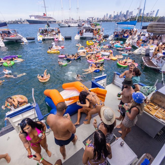 A group of people enjoying a sunny day on a boat, with many others floating on inflatables in the water. Boats and a city skyline are visible in the background, perfect for a Corporate boat event Sydney or even DJ boat hire Sydney to elevate the fun.