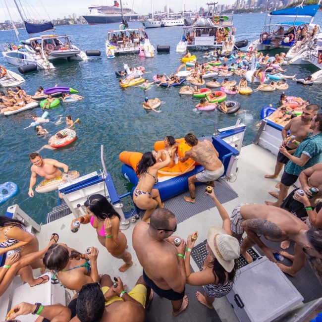 A lively floating party with people on boats and inflatables in a sunny marina. Attendees are swimming, socializing, and enjoying beverages. Numerous boats and yachts are visible in the background, highlighting the fun atmosphere of a DJ boat hire Sydney event.