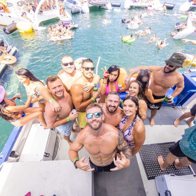 A group of people wearing swimwear gather on a boat, smiling and posing for a photo with a lively scene of other boats and people swimming in the background during a luxury yacht hire Sydney.