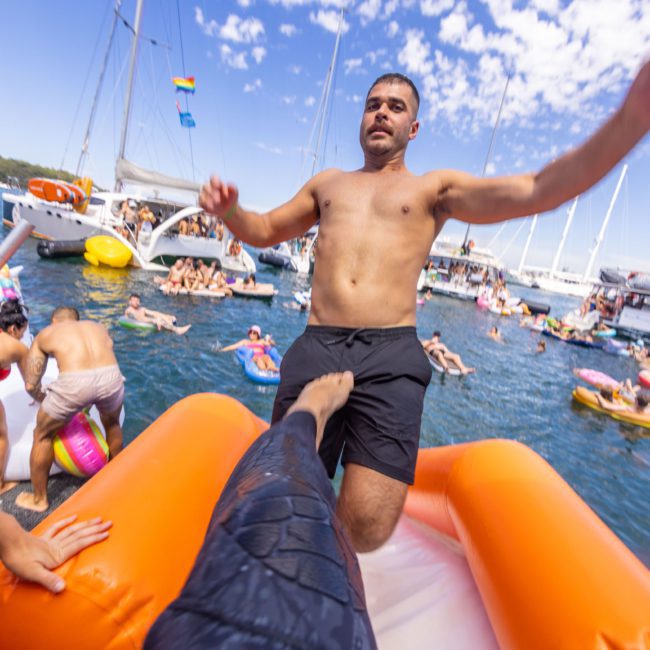 A person is balancing on an inflatable object in the water at a lively Sydney boat party hire event. Various floaties and boats are visible, highlighting the fun atmosphere. The clear sky with some clouds adds to the perfect setting for this memorable gathering.