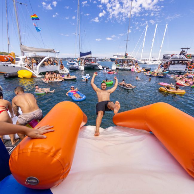 People enjoy a sunny day on a crowded body of water with inflatable toys, floating structures, and boats around them as a person slides down an inflatable slider into the water. Among the vessels is a luxury yacht hire Sydney, adding a touch of elegance to the lively scene.