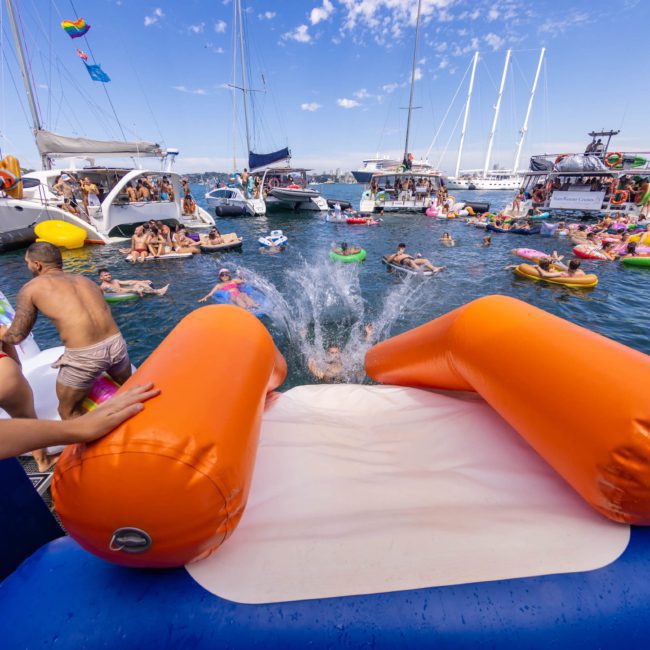 A crowd enjoys a summer party on boats and floats in the ocean, with someone sliding into the water from an inflatable slide. Surrounded by various watercraft and swimmers under a blue sky, this is the essence of a catamaran party Sydney.