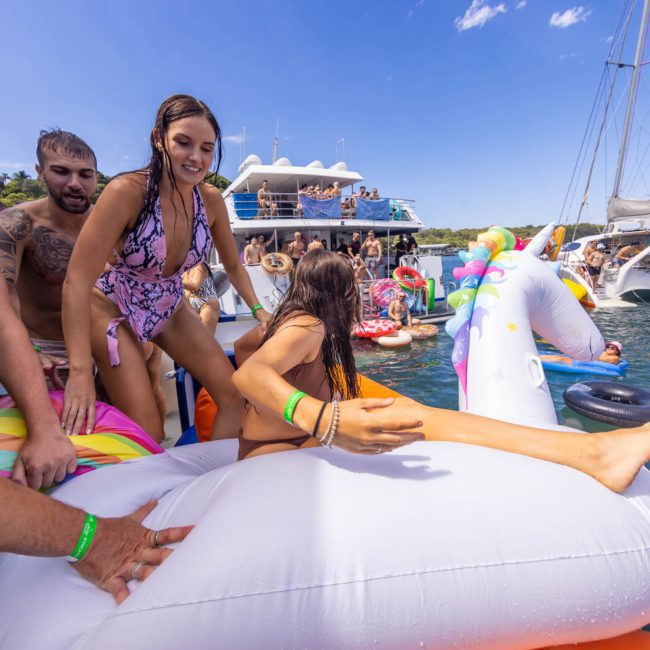A group of people in swimwear enjoys a sunny day on a large, white inflatable unicorn float in the water, with boats and more people in the background, making it an ideal scene for a Sydney boat party hire.