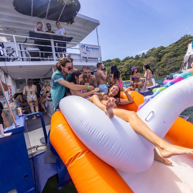 People on a boat and in the water are enjoying a sunny day. One person is sliding down an inflatable slide into the water, while others watch and interact nearby, making it a perfect setting for corporate boat events Sydney.
