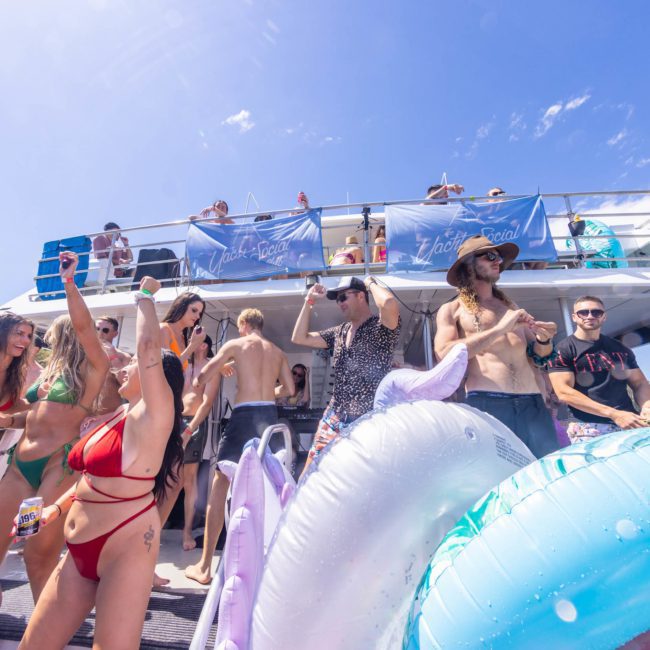 People in swimwear are enjoying a Sydney boat party hire on a yacht under a clear blue sky. Inflatable pool toys are in the foreground, and attendees are dancing, talking, and taking photos. It's a perfect setting for corporate boat events Sydney or simply having fun with friends under the sun.