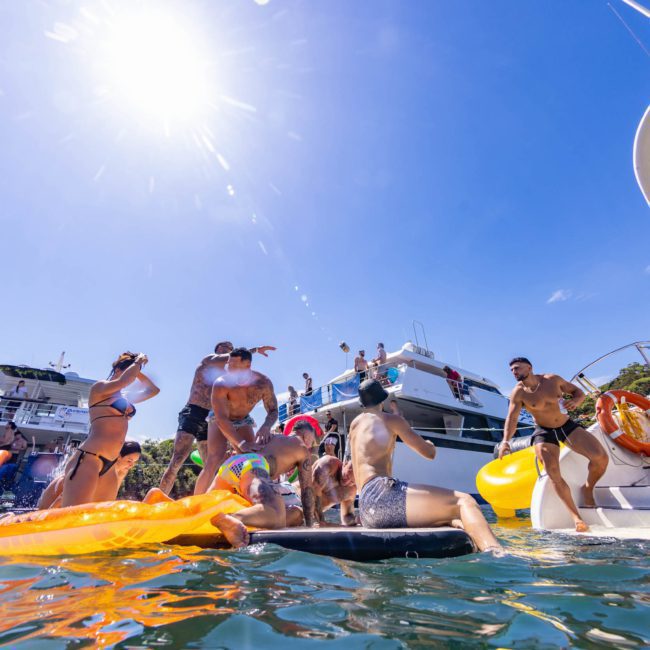 A group of people in swimsuits are enjoying a sunny day on the water, surrounded by boats and various inflatables. The bright sun shines overhead in a clear sky. It’s a perfect setting for a Sydney boat party hire, making the day unforgettable.