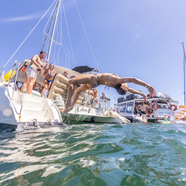 A person in a bikini jumps off a boat into the water, with several other people nearby on the boat. Multiple boats and a clear blue sky are in the background, capturing the essence of luxury yacht hire Sydney.