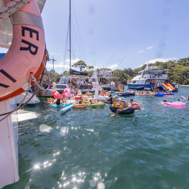 Boats docked on a sunny day with people enjoying the water on various inflatables nearby. In the background, there’s one boat with a visible life ring on the left side. It's an ideal scene for a Sydney boat party hire or corporate boat events in Sydney.
