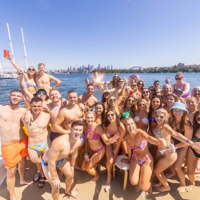 A large group of people in swimwear poses for a photo on a boat against a backdrop of water, city skyline, and other boats, showcasing the fun of corporate boat events Sydney.