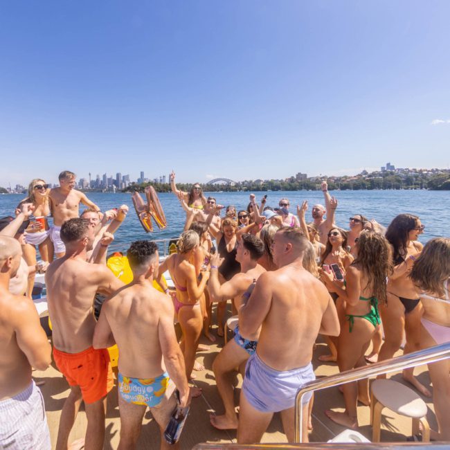 A group of people in swimwear are gathered on a luxury yacht hire in Sydney, enjoying a party under sunny skies. The background shows water, a city skyline, and several buildings on the shore.