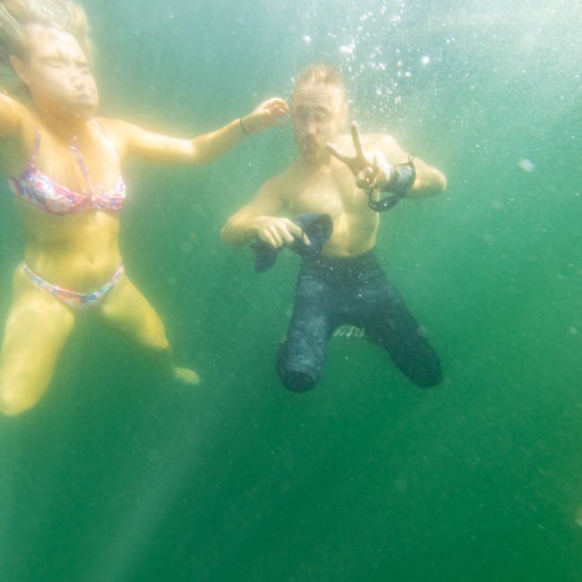 Two people underwater, one in a bikini and one in a wetsuit making a peace sign, with sunlight filtering through the green-tinged water; imagine this as part of an unforgettable private yacht charter on Sydney Harbour.