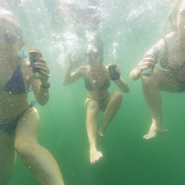 Three individuals in swimwear holding cans while underwater during a lively Catamaran party in Sydney.