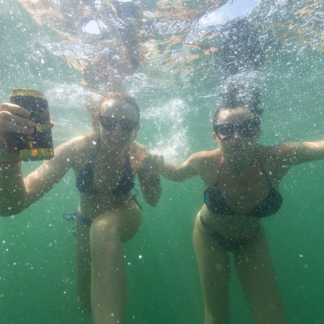 Two people underwater, both wearing swimsuits and sunglasses, enjoying a luxury yacht hire in Sydney, with one person holding a can.