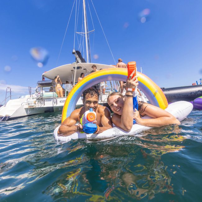 Two people float on an inflatable unicorn in the ocean, surrounded by boats and other people. They hold water guns and smile under a clear blue sky, enjoying what feels like a spontaneous Sydney boat party hire experience.