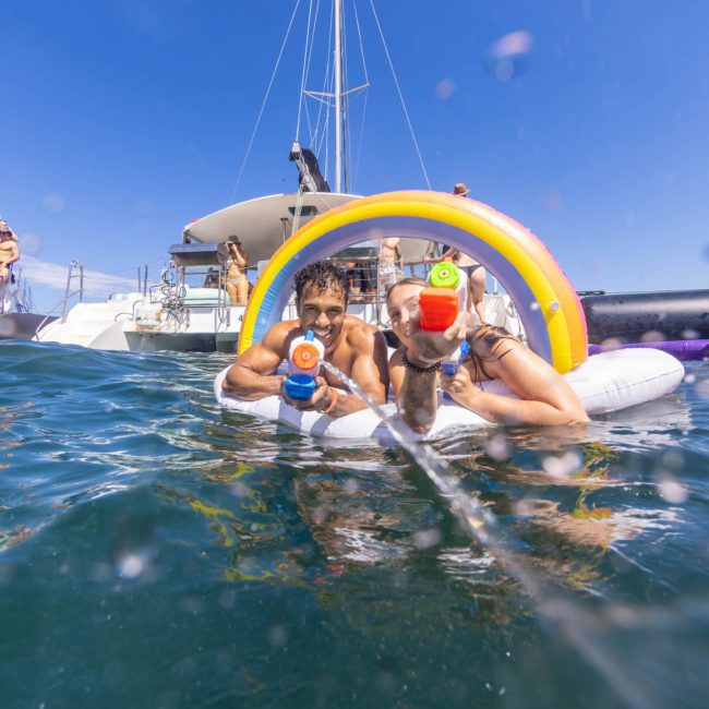 Two people on an inflatable raft with rainbow arches holding water guns in a body of water, enjoying the sun during a private yacht charter Sydney Harbour event. Boats and other people are visible in the background.