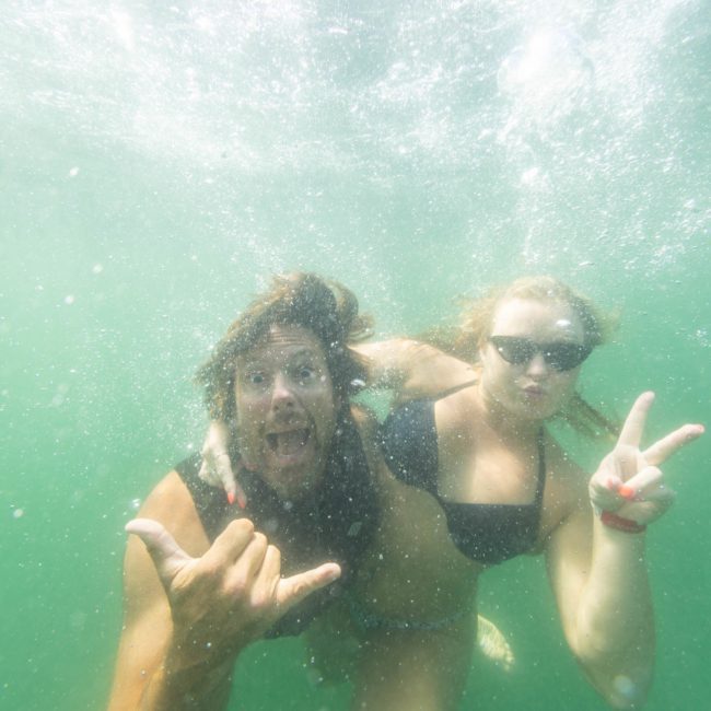 Two people underwater in swimwear making hand gestures and expressions. One person is making a "shaka" sign, and the other is showing a peace sign. Bubbles float around them, reminiscent of the fun vibes at a Sydney boat party hire.