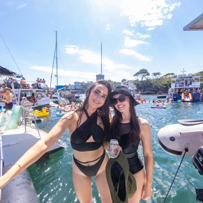 Two women in black swimsuits stand on a boat's deck, smiling at the camera. Surrounded by water and other boats, people enjoy a sunny day during a catamaran party in Sydney.