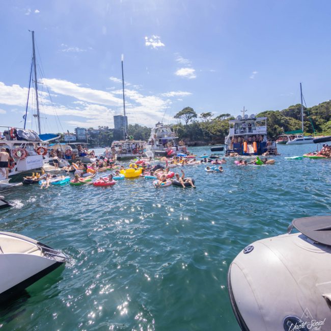Boaters anchored near a shore with people swimming and floating on inflatables in clear blue water under a sunny sky, enjoying the ultimate Sydney boat party hire experience.