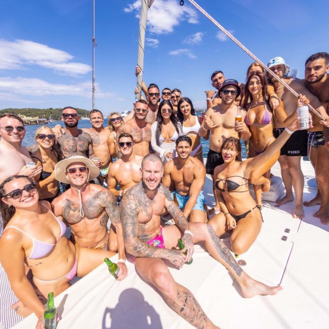 A large group of people in swimsuits are gathered on a catamaran under clear skies, smiling and posing for the camera. Some are holding drinks, and the water and shoreline are visible in the background.