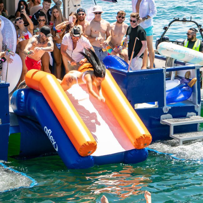 A group of people are enjoying a catamaran party in Sydney. One person is sliding down an inflatable slide into the water while others observe and cheer. Some are wearing swimsuits and sunglasses, soaking up the fun atmosphere of this vibrant Sydney boat party hire.