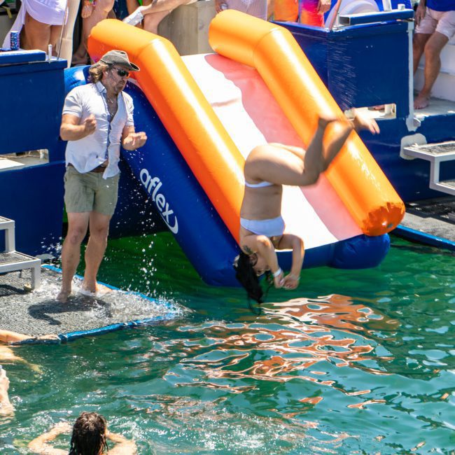 People enjoying a party on a floating platform with a water slide off a luxurious yacht hire in Sydney. A woman is mid-flip on the slide, and spectators are watching. Some people are in the water, and others are on the platform, making it perfect for corporate boat events in Sydney.