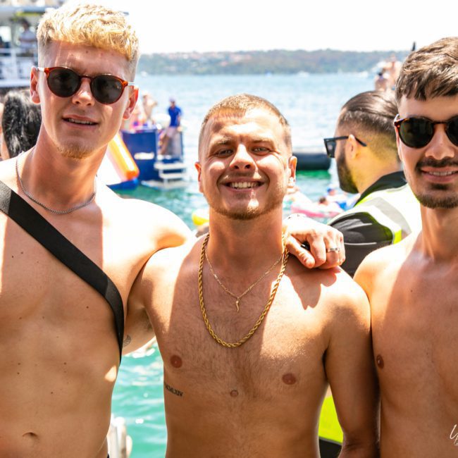 Three shirtless men wearing sunglasses, smiling and posing together near a waterfront during a private yacht charter Sydney Harbour. A boat and other people are visible in the background.