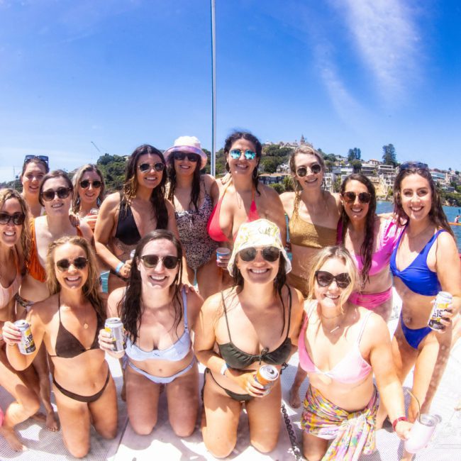 A group of people in swimsuits pose for a photo on a boat under a clear blue sky, some holding canned drinks, with water and a shoreline visible in the background during an elegant catamaran party Sydney.
