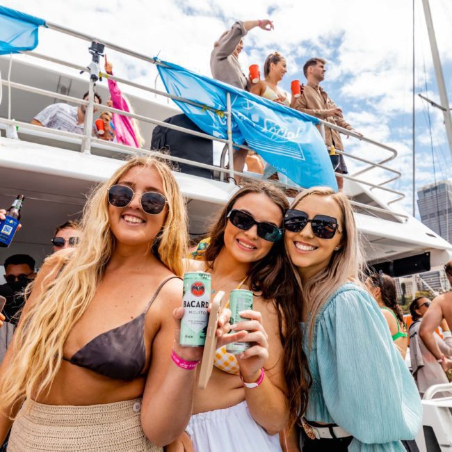 A group of three women with drinks smile for the camera on a boat. More people are visible on the upper deck, under blue flags, enjoying a sunny day during a Sydney boat party hire.