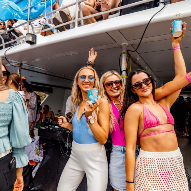 Three women pose and smile for the camera while holding drinks at a luxury yacht hire Sydney event, enjoying an outdoor party with multiple people on a multi-level boat.