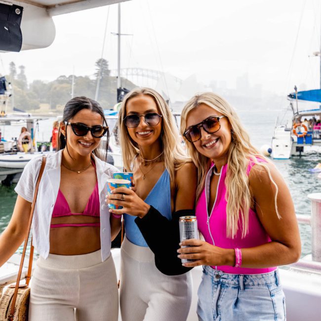 Three women wearing sunglasses and casual summer clothing pose and smile on a boat, with other boats and a bridge visible in the background, enjoying what looks like a perfect day for a private yacht charter Sydney Harbour.